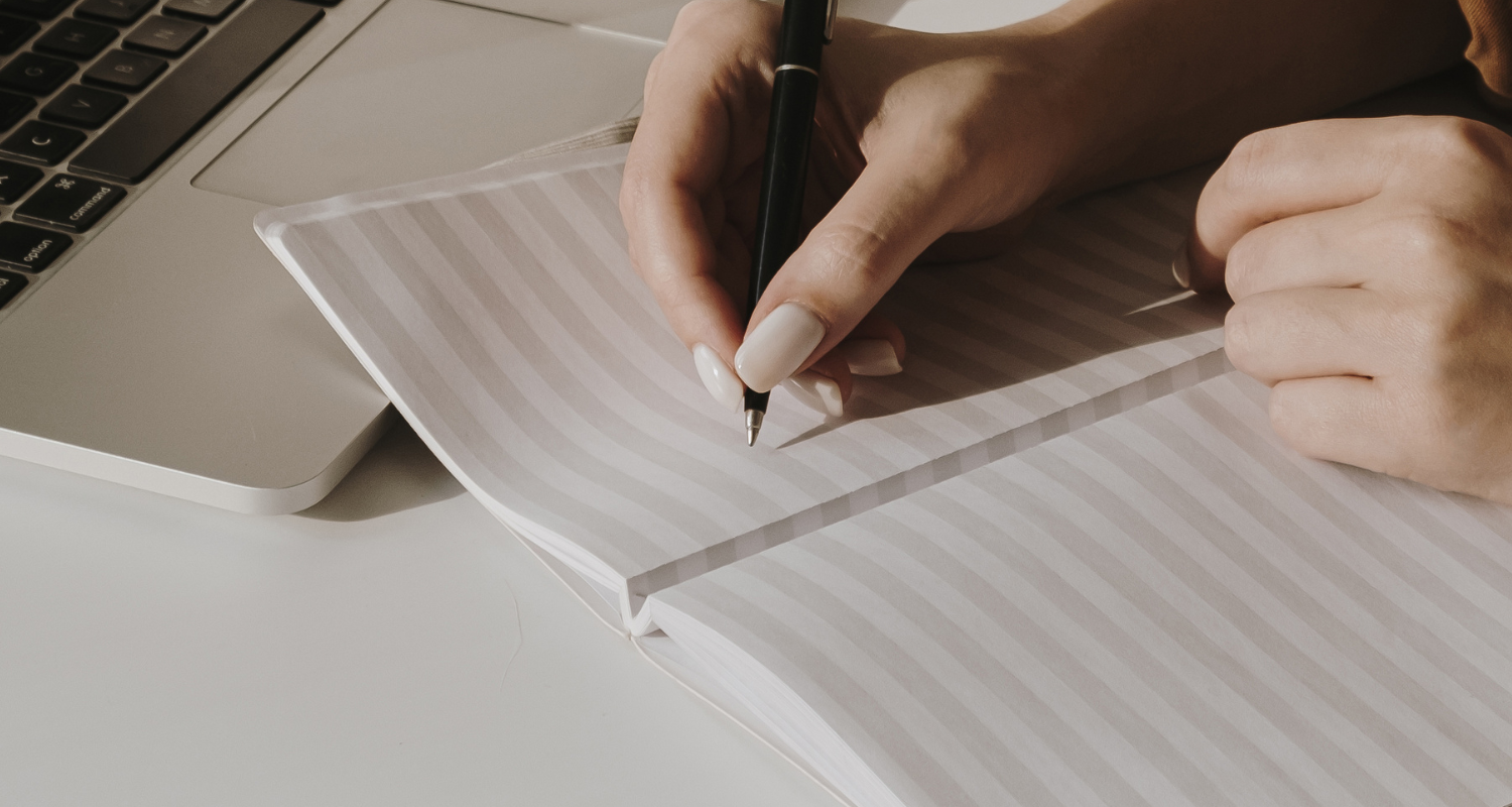 Woman writing in book with pencil at desk with laptop un front