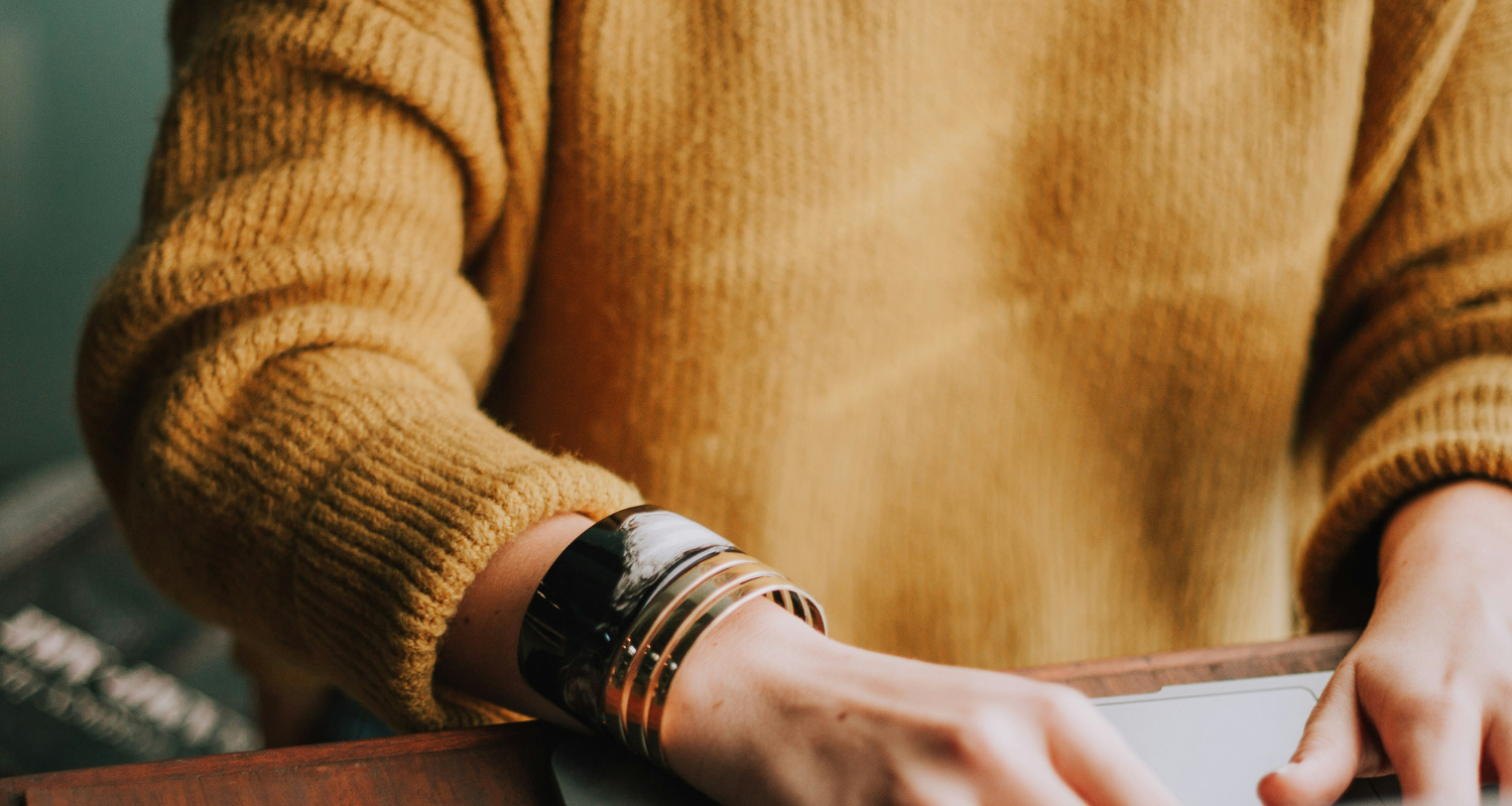 Woman sitting at table typing in yellow sweater and wearing gold bangles